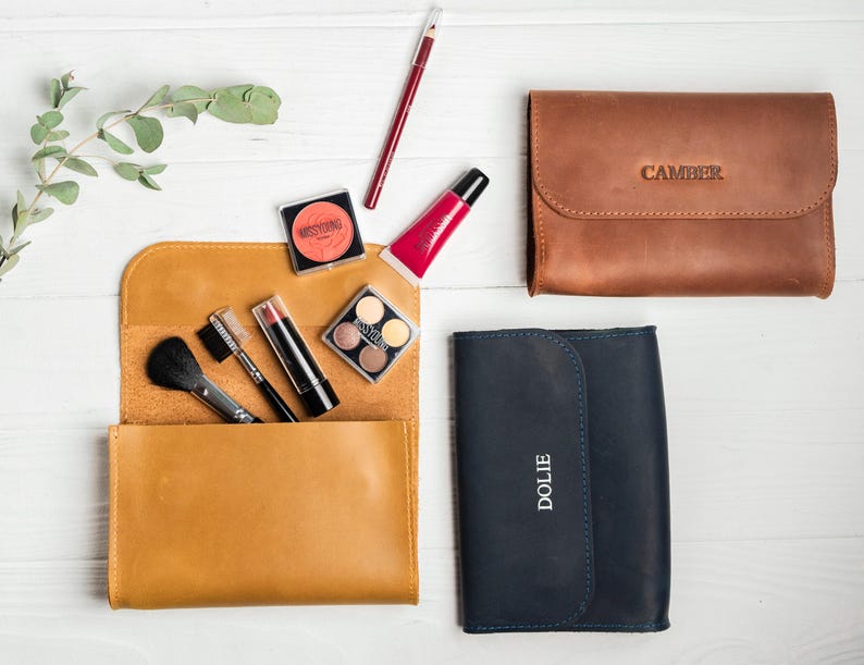 May include: An overhead shot of makeup and leather cosmetic bags. A mustard yellow leather bag is open, revealing makeup brushes, lipstick, and eyeshadow. A brown leather bag with "CAMBER" embossed on it and a navy blue bag with "DOLIE" are also present.
