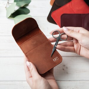 May include: A brown leather case with a flap closure and a small pocket on the front. The case is personalized with the initials "UC". A pair of small silver scissors is lying next to the case.