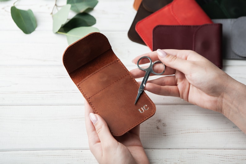 May include: A brown leather case with a flap closure and a small pocket on the front. The case is personalized with the initials "UC". A pair of small silver scissors is lying next to the case.