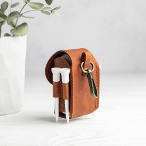 May include: A brown leather golf ball pouch featuring a flap closure, a brass-toned metal swivel clip, and two exterior loops holding white golf tees. The pouch is constructed with visible stitching along the edges and sides.