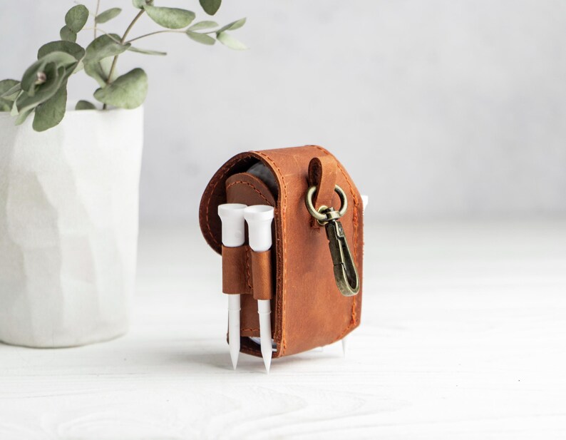 May include: A brown leather golf ball pouch featuring a flap closure, a brass-toned metal swivel clip, and two exterior loops holding white golf tees. The pouch is constructed with visible stitching along the edges and sides.