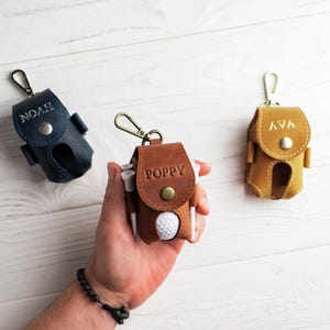 May include: Three personalized leather golf ball pouches in navy blue, brown, and tan. Each pouch has a snap closure and a metal clip. The brown pouch has the name "POPPY" embossed, while the navy and tan pouches have "NOAH" and "AVA" embossed.
