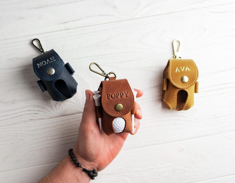 May include: Three personalized leather golf ball pouches in navy blue, brown, and tan. Each pouch has a snap closure and a metal clip. The brown pouch has the name "POPPY" embossed, while the navy and tan pouches have "NOAH" and "AVA" embossed.