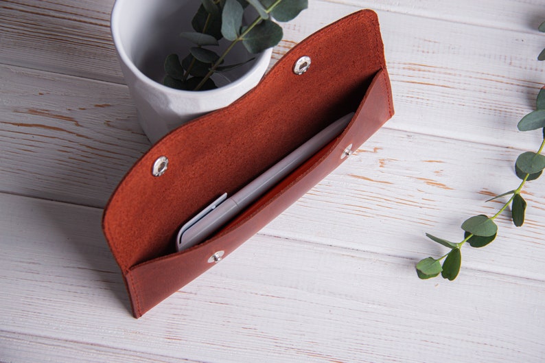 May include: A brown leather pencil case with a silver snap closure. The case is open and a silver pen is inside.