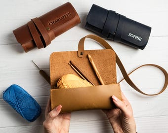 May include: A tan leather crochet hook case with a handle, open to reveal crochet hooks and yellow yarn. Two rolled leather cases, one brown with "CHARLOTTE" and one navy with "SOPHIE" are also shown. A blue ball of yarn is on the left.