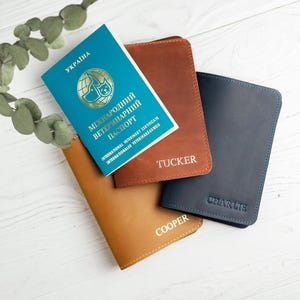 Könnte beinhalten: Eine Auswahl an Leder-Passhüllen in Braun, Beige und Marineblau, personalisiert mit Namen. Ein blau-goldenes ukrainisches internationales Veterinärzertifikat liegt obenauf. Die Artikel sind auf einer weißen Holzoberfläche mit grünem Laub angeordnet.