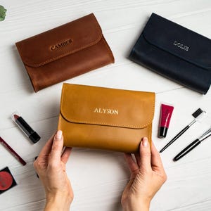 May include: Three leather cosmetic cases in brown, navy, and tan, each with a name embossed on the front. Makeup brushes, lipstick, and other cosmetic items are arranged around the cases on a white surface.