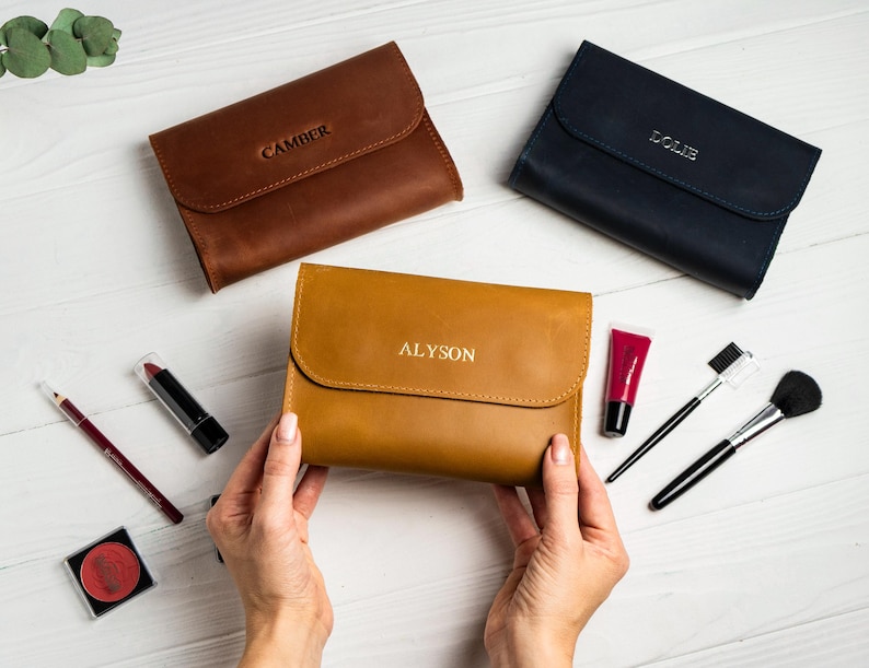 May include: Three leather cosmetic cases in brown, navy, and tan, each with a name embossed on the front. Makeup brushes, lipstick, and other cosmetic items are arranged around the cases on a white surface.