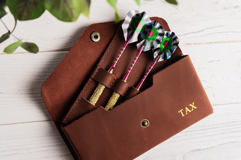 May include: A brown leather case with three darts inside. The case has a gold snap closure and the word "TAX" is embossed on the front.