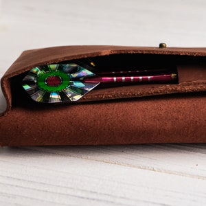 May include: A brown leather dart case with a single dart inside. The dart has a green and iridescent fletching.