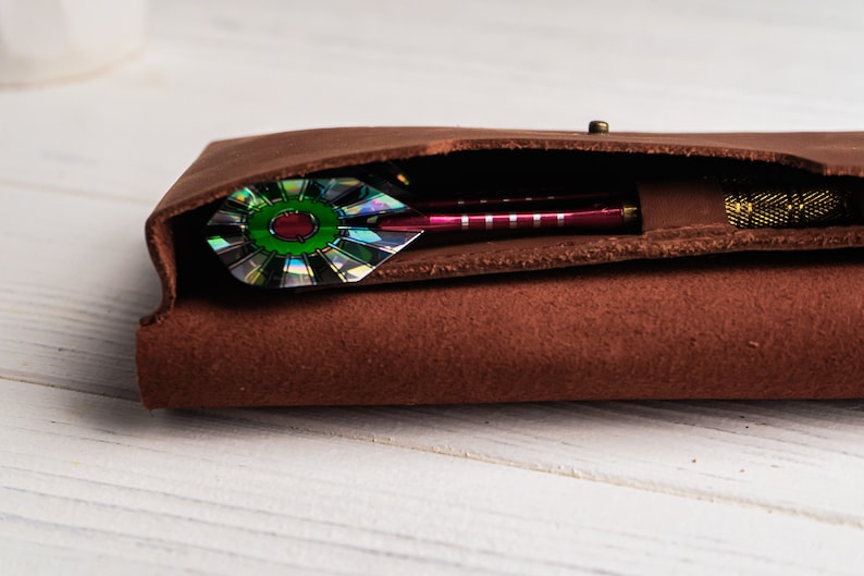 May include: A brown leather dart case with a single dart inside. The dart has a green and iridescent fletching.