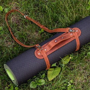 May include: A rolled-up black yoga mat with a brown leather carrying strap and shoulder strap. The mat is lying on green grass. The leather strap has silver-colored metal hardware.