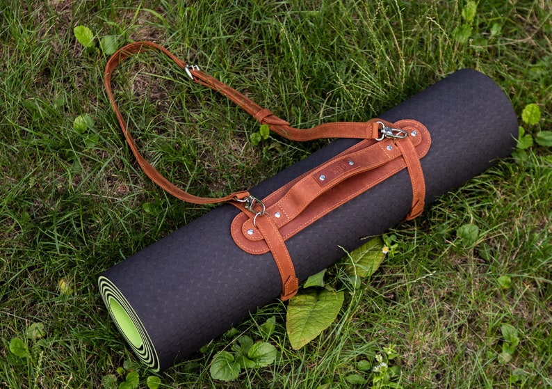 May include: A rolled-up black yoga mat with a brown leather carrying strap and shoulder strap. The mat is lying on green grass. The leather strap has silver-colored metal hardware.