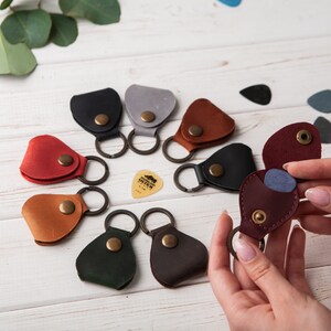 May include: Assortment of leather guitar pick holders with keychains in various colors, including red, brown, black, green, and gray. Each holder features a snap closure and a small pick inside.