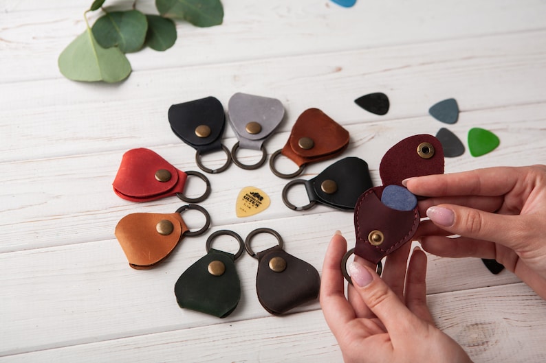 May include: Assortment of leather guitar pick holders with keychains in various colors, including red, brown, black, green, and gray. Each holder features a snap closure and a small pick inside.