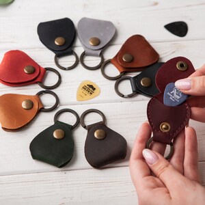 May include: A collection of leather guitar pick holders with key rings. The holders are in various colors, including brown, red, green, and blue. A pick is being removed from a burgundy holder.