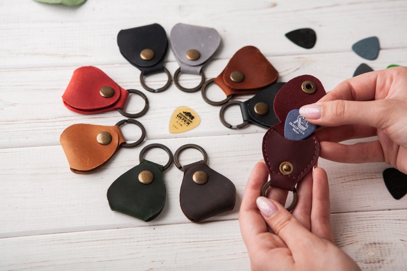 May include: A collection of leather guitar pick holders with key rings. The holders are in various colors, including brown, red, green, and blue. A pick is being removed from a burgundy holder.