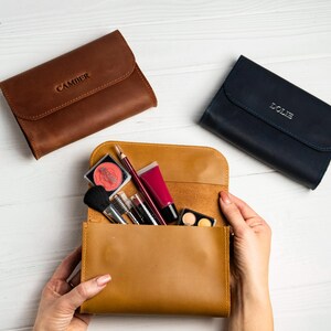 May include: Three leather cosmetic bags in brown, tan, and navy blue. The brown bag has the name "CAMBER" embossed on the flap. The tan bag is open, revealing makeup brushes, lipsticks, and eyeshadow. The navy blue bag has the name "LOLIE" embossed on the flap.