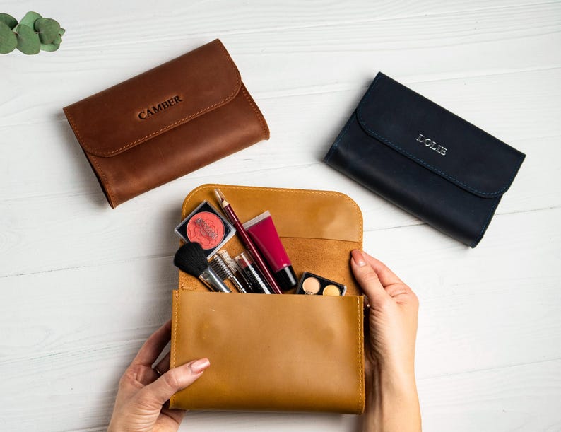 May include: Three leather cosmetic bags in brown, tan, and navy blue. The brown bag has the name "CAMBER" embossed on the flap. The tan bag is open, revealing makeup brushes, lipsticks, and eyeshadow. The navy blue bag has the name "LOLIE" embossed on the flap.