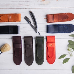 Op de afbeelding: Een assortiment leren scheermesetuis in diverse kleuren, waaronder bruin, zwart en blauw, met een scheermes en een scheerkwast. Sommige etuis hebben initialen. De items zijn gerangschikt op een wit houten oppervlak.