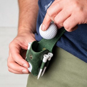 May include: A dark green leather golf ball holder with white golf tees. A person is placing a white golf ball into the holder. The holder is attached to a belt loop. The person is wearing a navy blue shirt and khaki pants.