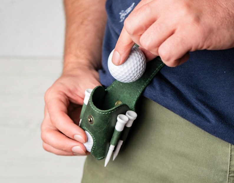 May include: A dark green leather golf ball holder with white golf tees. A person is placing a white golf ball into the holder. The holder is attached to a belt loop. The person is wearing a navy blue shirt and khaki pants.