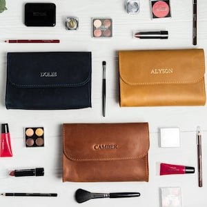May include: An overhead shot of makeup products and accessories, including three leather cosmetic bags with embossed names: "Dolie", "Alyson", and "Camber". The image also features eyeshadow palettes, lipsticks, brushes, and other beauty items, all arranged on a white surface.