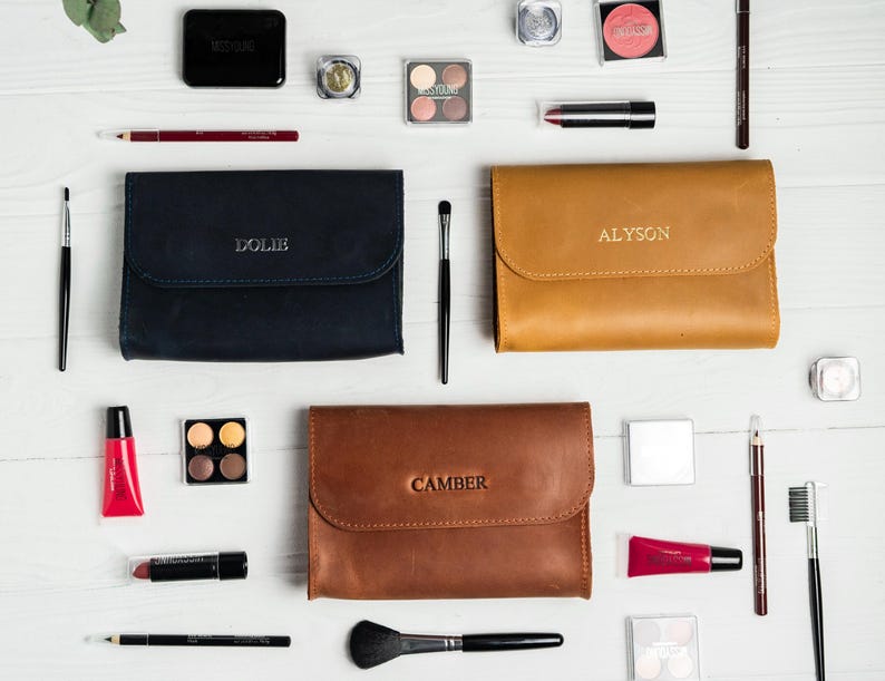 May include: An overhead shot of makeup products and accessories, including three leather cosmetic bags with embossed names: "Dolie", "Alyson", and "Camber". The image also features eyeshadow palettes, lipsticks, brushes, and other beauty items, all arranged on a white surface.