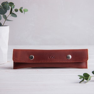 May include: A brown leather pencil case with silver snaps and the initials "OM" embossed on the front.