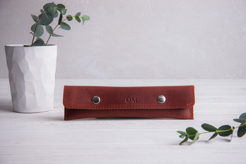 May include: A brown leather pencil case with silver snaps and the initials "OM" embossed on the front.
