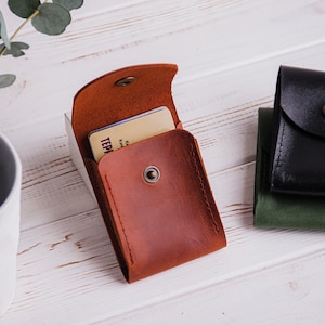 Pu&ograve; includere: Un porta carte in pelle marrone con chiusura a scatto, aperto per rivelare una carta. Sono visibili anche due porta carte in pelle aggiuntivi in nero e verde. Gli articoli sono su una superficie di legno bianco.