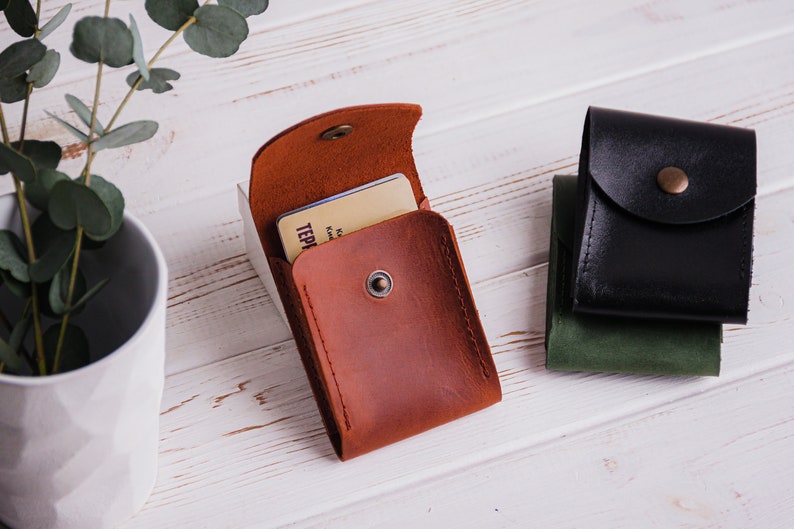 Pu&ograve; includere: Un porta carte in pelle marrone con chiusura a scatto, aperto per rivelare una carta. Sono visibili anche due porta carte in pelle aggiuntivi in nero e verde. Gli articoli sono su una superficie di legno bianco.