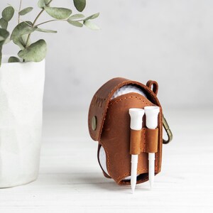 May include: A brown leather golf ball holder with two white golf tees attached. The holder has a snap closure and a metal clip. A white golf ball is visible inside. A white vase with green leaves is in the background.