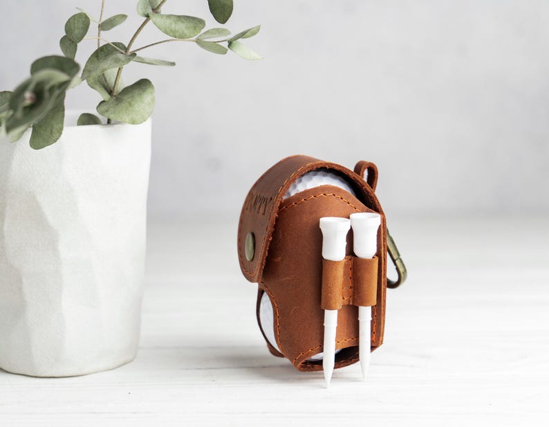 May include: A brown leather golf ball holder with two white golf tees attached. The holder has a snap closure and a metal clip. A white golf ball is visible inside. A white vase with green leaves is in the background.