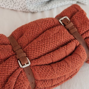 May include: A rolled-up, rust-colored knitted blanket secured with two brown leather straps and silver buckles. The blanket has a textured pattern and is neatly folded. The background is a soft, neutral color.