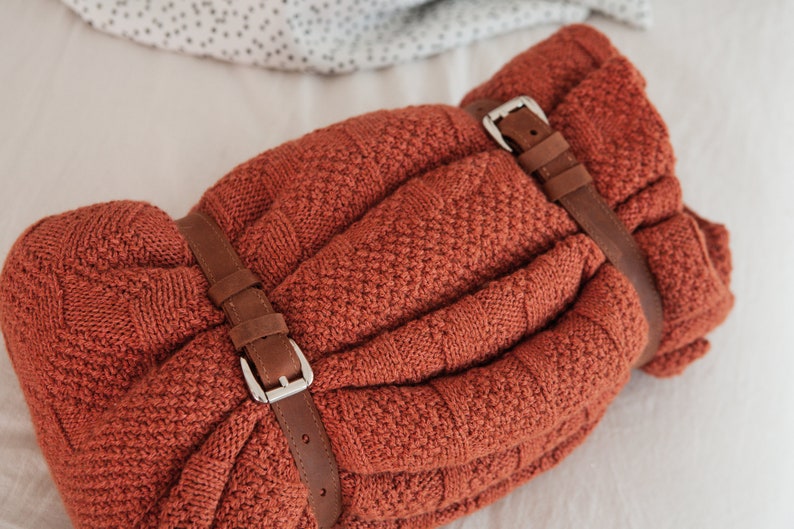 May include: A rolled-up, rust-colored knitted blanket secured with two brown leather straps and silver buckles. The blanket has a textured pattern and is neatly folded. The background is a soft, neutral color.