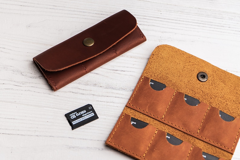 K&ouml;nnte beinhalten: Zwei braune Lederetuis zum Aufbewahren von Speicherkarten. Das gr&ouml;&szlig;ere Etui hat sechs Steckpl&auml;tze f&uuml;r Speicherkarten. Das kleinere Etui hat einen Steckplatz f&uuml;r eine Speicherkarte. Neben den Etuis ist eine Micro-SD-Karte abgebildet.