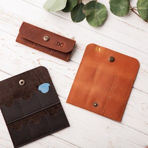 May include: Three leather keychains with different designs. The keychain on the left has six slots and is brown. The keychain in the middle is a slim brown case with the initials "DC" embossed on it. The keychain on the right is a brown case with six slots and the initials "KA" embossed on it.