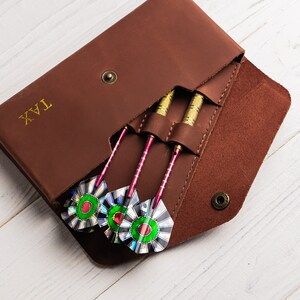 May include: Brown leather dart case with the word "TAX" embossed on the front. The case is open, revealing three darts with green and iridescent flighted tips.