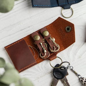 Könnte beinhalten: Ein braunes Schlüsseletui aus Leder mit einer Umschlagklappe und einem Druckknopfverschluss. Das Innere enthält zwei Lederriemen mit Druckknöpfen zur Befestigung von Schmuck, darunter eine goldene Kette und ein Ring, an einem Metallschlüsselring.