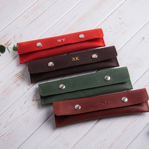 May include: Five leather pencil cases in different colors, each with a different monogram. The cases are arranged in a stack, with the red case on top and the brown case on the bottom. The monograms are WY, XK, OM, and two others.