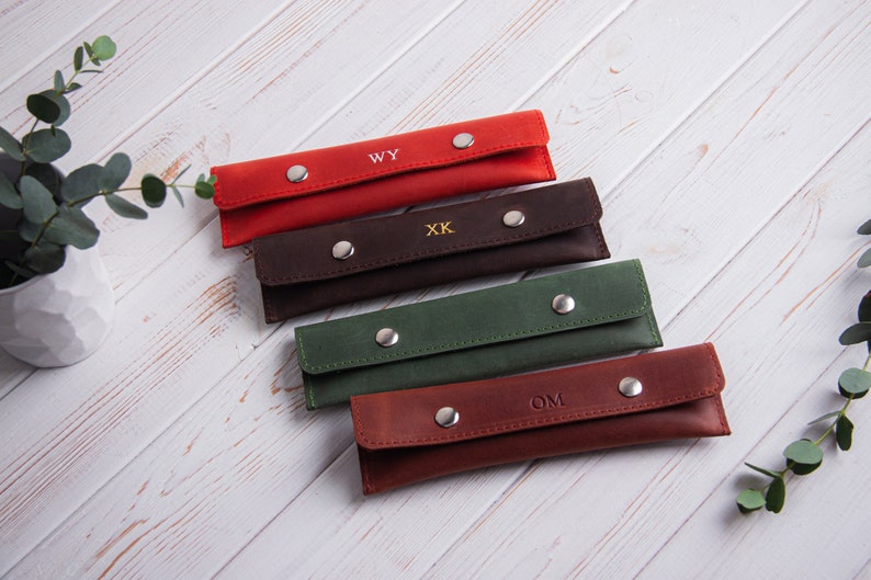 May include: Five leather pencil cases in different colors, each with a different monogram. The cases are arranged in a stack, with the red case on top and the brown case on the bottom. The monograms are WY, XK, OM, and two others.