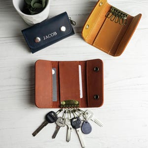 May include: Leather key organizers in navy blue, tan, and brown, with keys attached. One navy blue organizer is embossed with the name "JACOB". The organizers are open, displaying key rings and card slots. A small white pot with greenery is in the background.