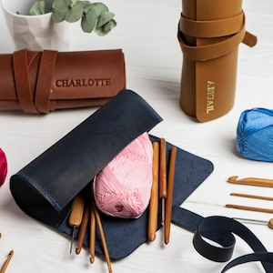 May include: A collection of crochet tools and supplies, including a dark blue leather crochet hook case with a pink yarn ball and wooden hooks. Other items include brown leather cases with the names "CHARLOTTE" and "ABIGAIL" embossed on them, and balls of red and blue yarn.