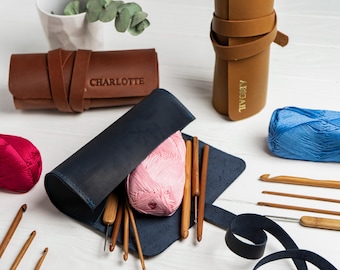 Può includere: Una collezione di strumenti e materiali per l'uncinetto. Un astuccio per uncinetti in pelle blu scuro contiene un gomitolo di lana rosa e uncinetti in legno. Sono presenti anche astucci in pelle marrone con i nomi "CHARLOTTE" e "ABIGAIL" in rilievo e filati rosso e blu.