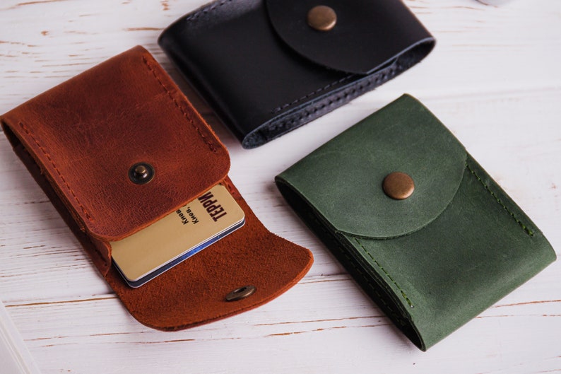 Pu&ograve; includere: Tre porta carte in pelle marrone, nera e verde, ciascuno con chiusura a scatto. Il porta carte marrone &egrave; aperto, rivelando una carta con testo. I porta carte sono su una superficie di legno bianco.
