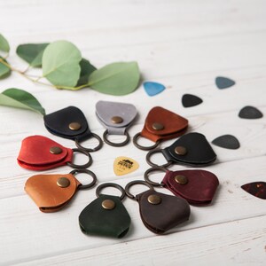May include: A circle of ten leather guitar pick holders with key rings. Each holder is a different color: red, brown, black, gray, green, dark brown, burgundy, light brown, and blue. A single guitar pick is in the center of the circle.