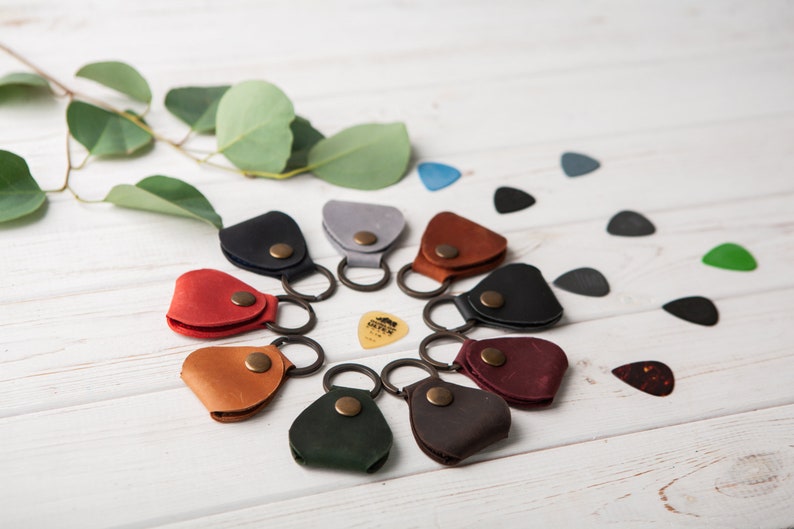 May include: A circle of ten leather guitar pick holders with key rings. Each holder is a different color: red, brown, black, gray, green, dark brown, burgundy, light brown, and blue. A single guitar pick is in the center of the circle.