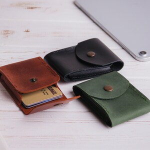 Pu&ograve; includere: Tre porta carte in pelle marrone, nera e verde, ciascuno con chiusura a scatto. Il porta carte marrone &egrave; aperto, rivelando diverse carte. Un tablet argentato &egrave; sullo sfondo, appoggiato su una superficie di legno bianco.