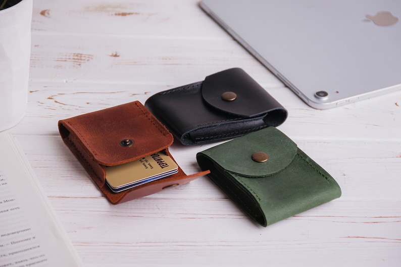 Pu&ograve; includere: Tre porta carte in pelle marrone, nera e verde, ciascuno con chiusura a scatto. Il porta carte marrone &egrave; aperto, rivelando diverse carte. Un tablet argentato &egrave; sullo sfondo, appoggiato su una superficie di legno bianco.